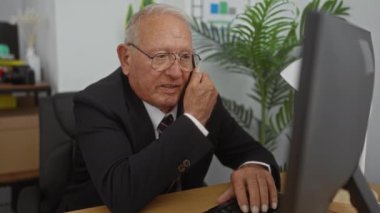 Yaşlı, gri saçlı, takım elbiseli beyaz bir adam ofis ortamında masa başında çalışırken telefonla konuşuyor..