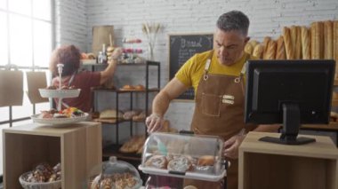 Pastanede çalışan bir adam pastaları düzenlerken arka plandaki bir kadın içeride raflarda ekmek düzenliyor..