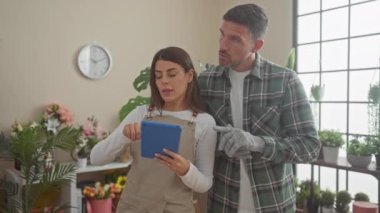 Bir erkek ve bir kadın çiçekçi, çeşitli bitkilerle dolu kapalı çiçek dükkanlarını yönetmek için bir tablet kullanıyorlar..