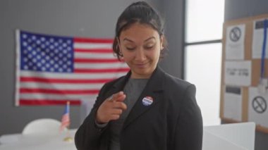 Neşeli, genç İspanyol bir kadın arka planda Amerikan bayrağı olan bir oy kullanma merkezindeki 