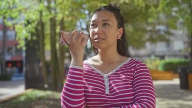 Genç bir İspanyol kadın, güneşli, yapraklı bir şehir parkında, pembe çizgili bir bluzla akıllı telefonuyla konuşuyor..