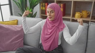 Kendine güvenen, tesettürlü genç İspanyol kadın, gururla oturma odasında kendini işaret ediyor. Zaferi kutluyoruz. Kanepede oturup, mutlulukla gülümsüyoruz..