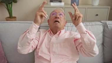 Gözlüklü, yaşlı, beyaz bir adam iki eli havada, oturma odasındaki bir kanepede oturuyor..