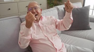 Son sınıf öğrencisi gözlüklü ve pembe gömlekli bir adam modern bir oturma odasında koltukta otururken kollarını kaldırarak kutlama yapıyor..