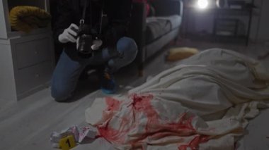 Kel bir adam, kapalı bir suç mahallinde kan lekeleri ve üstü örtülü bir cesetle kanıt fotoğrafı çekiyor..
