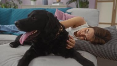 Genç bir kadın, siyah labrador köpeğiyle rahat bir oturma odasında kanepede uzanıyor ve birlikte rahat bir anın tadını çıkarıyor..