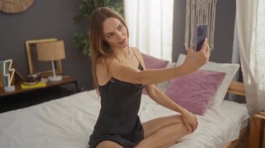 Güzel, genç, sarışın bir kadın, modern dekorlu, rahat bir yatak odasında otururken selfie çekiyor..