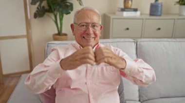 Oturma odasında, rahat bir evde, kanepede otururken elleriyle kalp şekli yapan yaşlı, beyaz bir adam..