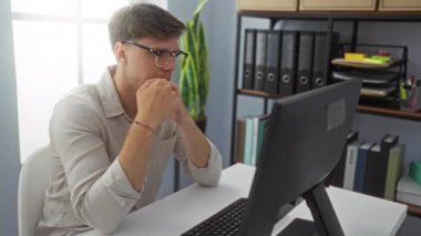 Ofiste çalışan genç bir adam bilgisayar ve bitkilerle dolu bir masada oturuyor, gözlük takıyor ve stresli ya da yorgun görünüyor..