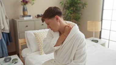 Beyaz bornozlu genç bir adam bej duvarlı bir yatak odasında bir yatakta oturuyor ve boynunu acı içinde tutuyor.