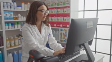 Beyaz önlüklü ve gözlüklü genç bir İspanyol kadın eczanedeki bir bilgisayarda çeşitli ürünlerle dolu raflarla çalışıyor..