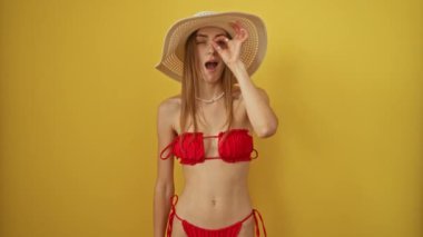 Yaz bikinili ve şapkalı kadın sarı arka planda şaşırmış bir ifadeyle parmaklarının arasından bakıyor.