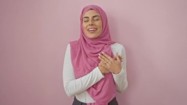 İspanyol kadın tesettürlü, elleri göğsünde, gönülden şükranlarını ifade ediyor sağlık mücadeleleri arasında, pembe duvarın üzerinde izole bir şekilde.