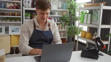 Genç adam dekoratif eşyalar, bitkiler ve paketlerle çevrili bir ev dekorasyonu mağazasında dizüstü bilgisayarda çalışıyor.