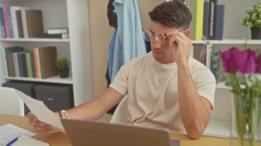 Düşünceli bir genç adam, evde kitap ve dizüstü bilgisayarla çevrili bir belgeyi incelerken gözlüklerini çıkarıyor..