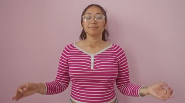 Çizgili tişörtlü genç İspanyol kadın Zen 'i ayakta, ayakta, gözleri kapalı meditasyon yaparken ve pembe izole edilmiş arka plan üzerinde yoga hareketi yaparken buluyor.