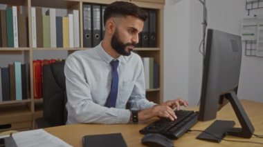 Kitaplar ve klasörlerle dolu bir ofis ortamında bilgisayar ve tablet üzerinde çalışan sakallı genç bir İspanyol.