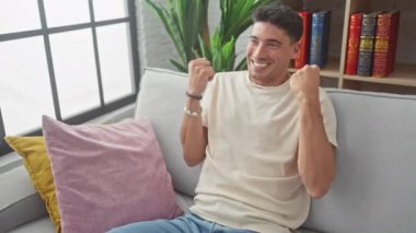 Evdeki koltukta oturan heyecanlı İspanyol genç adam muzaffer bir sevinçle oturuyor, kolları mutlu bir başarı kutlamasıyla yükseliyor. Evet! büyük kazanç!