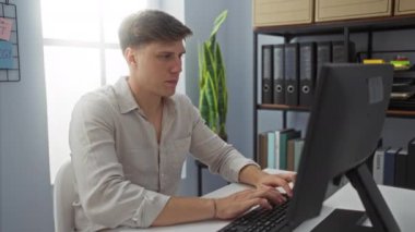 Ofiste çalışan beyaz bir genç, arka planda bitki ve kitaplık olan bir masada otururken bilgisayarına odaklanmış.