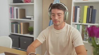 Ofiste gülümseyen kulaklıklı İspanyol adam, kitaplık, gözlük, günlük, dinleme, iç mekan, genç, çekici, yetişkin, erkek.