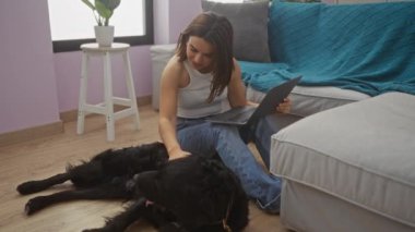 Genç bir kadın siyah bir labrador ile oturma odasında dizüstü bilgisayar kullanarak rahat bir ortam yaratıyor..