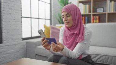 Başörtüsü içinde gülümseyen bir kadın, elinde bir kredi kartı ve akıllı telefon tutuyor. Bu da samimi bir oturma odasında online alışveriş yaptığını gösteriyor..