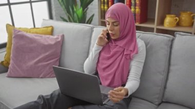 Modern bir oturma odasında pembe tesettürlü, telefon görüşmesi ve dizüstü bilgisayarlı bir kadın..