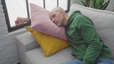 Yeşil kazaklı kel bir adam modern oturma odasındaki renkli yastıklarla kanepede uyuyor..
