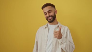 İspanyol adam gülümsüyor ve başparmak işareti yapıyor. İzole edilmiş sarı bir arka plana karşı duruyor. Kendine güvenen ve pozitif bir tavır sergiliyor..