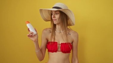 Bikinili bir kadın elinde güneş kremi tutarak gülümsüyor ve sarı arka planda başparmağıyla işaret ediyor.