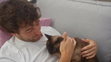 Sevecen bir adam, sıcak bir oturma odasında siyam kedisine sarılıyor..