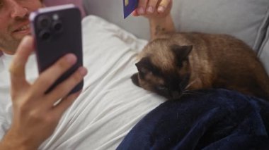 Adamın biri kredi kartıyla selfie çekiyor ve siyam kedisi evde kanepede uzanıyor..