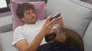 Rahat genç bir adam, rahat bir oturma odasında akıllı telefon kullanırken siyam kedisiyle kanepede uzanıyor..