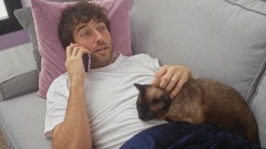 Gülümseyen bir adam telefonda kedi beslerken rahat mobilyalarla rahat bir ev ortamını resmediyor..