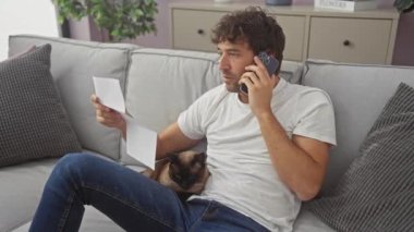 Adamın biri bir belgeyi incelerken telefonda konuşuyor. Yanında da evdeki kanepede oturan siyam kedisi var..