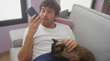 Genç İspanyol bir adam, oturma odasında siyam kedisiyle kanepede yatarken akıllı telefonuyla bir sesli mesaj kaydediyor..