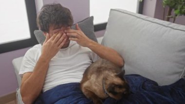 Genç bir İspanyol adam, oturma odasındaki kanepede kedinin kucağında otururken burnunu ovalıyor. Ev ortamında alerjisi olabileceğini öne sürüyor..