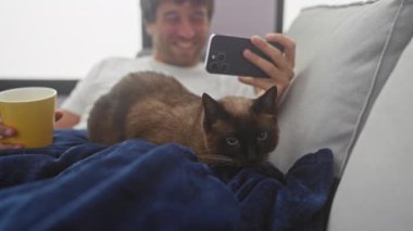 Gülümseyen bir adam elinde bir telefon ve bir kupa, bir kanepede siyam kedisiyle uzanıyor..