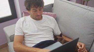 Beyaz tişörtlü bir adam dizüstü bilgisayarı ve kapalı koltukta uyuyan bir kediyle uzanıyor.