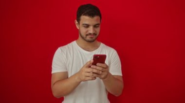 Genç İspanyol adam mutlu ve gülümserken parmaklarıyla akıllı telefonu işaret ederek izole edilmiş kırmızı arka planı teslim ediyor.