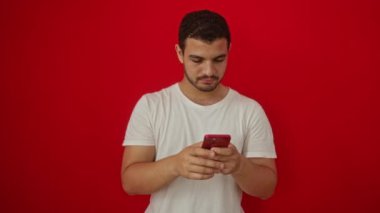 İspanyol asıllı genç adam tek eliyle gülümseyip yüzünü gösterirken kırmızı arka plan üzerinde akıllı telefon kullanarak mutluluk ve kendine güven yayıyor.