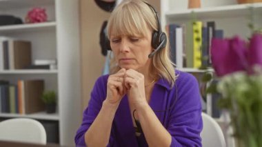 Mor kazaklı dalgın sarışın bir kadın ev ofisinde beyaz bir masa ve kitap rafı olan bir kulaklık kullanıyor..