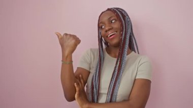 Neşeli Afro-Amerikalı kadın yukarıyı işaret ediyor, pembe bir zeminde yüzsüz bir gülümseme sergiliyor, mutlu bir jest sergiliyor, örgüler ve havalı bir tişörtle göze çarpıyor!