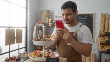 Fırında genç bir adam kırmızı telefonlu glütensiz pastaların fotoğrafını çekiyor. Etrafı taze ekmek ve pastalarla çevrili..