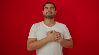 Beyaz tişört giyen genç İspanyol adam elleri göğsünde duruyor, izole edilmiş kırmızı arka planda minnet dolu bir jest yapıyor.