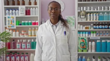 Beyaz laboratuvar önlüğü giyen Afrikalı kadın eczacı ürünlerle dolu bir eczanede güvenle duruyor..