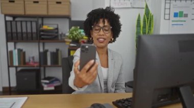 Ofisteki genç Afro-Amerikalı kadın telefonuyla selfie çekerken kapalı bir iş yerinde rafları ve ofis malzemeleriyle bir masada oturuyordu..