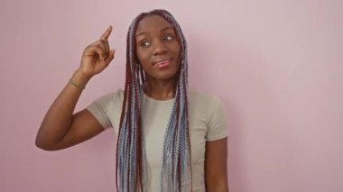 Pembe izole edilmiş arka planda duran örgülü neşeli Afrikalı Amerikalı kadın. Mutlu bir şekilde parmağınla işaret ederek harika bir fikir ya da iyi bir anı olduğunu gösterir.