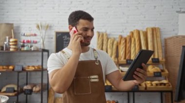 Ekmek ve hamur işleriyle çevrili, tablet kullanırken telefonda konuşan İspanyol bir adam.
