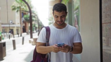 Genç İspanyol bir adam akıllı telefon kullanarak şehir caddesinde duruyor ve güneşli bir günde sırtüstü gülerek ve açık havada eğlenirken sırt çantası taşıyor.